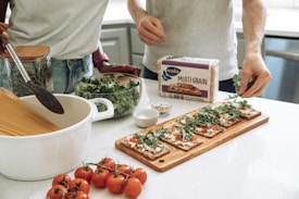 Two people are preparing a meal in a kitchen. There is a pot with dry spaghetti, a jar with herbs, and a bowl of greens on the counter. A package of multigrain crispbread is visible, along with a cutting board topped with crispbread, tomato sauce, cheese, and arugula. A bunch of tomatoes is placed in the foreground.