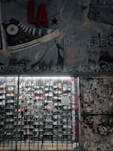 A street art mural with a large sneaker illustration and various graffiti tags covers a wall. There is a metal gate in front of a store, with visible shoe racks behind it. The wall to the right is plastered with numerous stickers and posters. The image has an urban, gritty aesthetic.