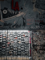 A street art mural with a large sneaker illustration and various graffiti tags covers a wall. There is a metal gate in front of a store, with visible shoe racks behind it. The wall to the right is plastered with numerous stickers and posters. The image has an urban, gritty aesthetic.