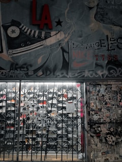 A street art mural with a large sneaker illustration and various graffiti tags covers a wall. There is a metal gate in front of a store, with visible shoe racks behind it. The wall to the right is plastered with numerous stickers and posters. The image has an urban, gritty aesthetic.