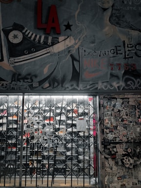 A street art mural with a large sneaker illustration and various graffiti tags covers a wall. There is a metal gate in front of a store, with visible shoe racks behind it. The wall to the right is plastered with numerous stickers and posters. The image has an urban, gritty aesthetic.