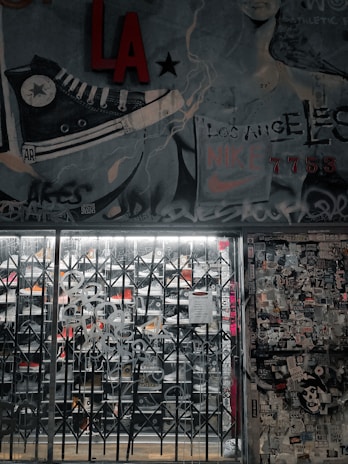 A street art mural with a large sneaker illustration and various graffiti tags covers a wall. There is a metal gate in front of a store, with visible shoe racks behind it. The wall to the right is plastered with numerous stickers and posters. The image has an urban, gritty aesthetic.