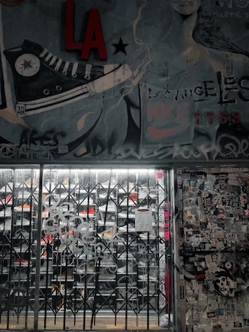 A street art mural with a large sneaker illustration and various graffiti tags covers a wall. There is a metal gate in front of a store, with visible shoe racks behind it. The wall to the right is plastered with numerous stickers and posters. The image has an urban, gritty aesthetic.