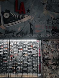 A street art mural with a large sneaker illustration and various graffiti tags covers a wall. There is a metal gate in front of a store, with visible shoe racks behind it. The wall to the right is plastered with numerous stickers and posters. The image has an urban, gritty aesthetic.