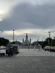 A cloudy sky dominates the background, with a large cathedral centered in the distance. The foreground features a street musician playing a keyboard, set up with equipment on a trolley. Several people are walking in the plaza leading to the cathedral, with trees and lampposts lining the path.