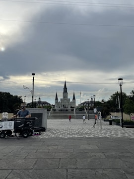 A cloudy sky dominates the background, with a large cathedral centered in the distance. The foreground features a street musician playing a keyboard, set up with equipment on a trolley. Several people are walking in the plaza leading to the cathedral, with trees and lampposts lining the path.