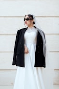 A person wearing a white wedding dress paired with a black jacket, sunglasses, and a veil, against a light-colored background. The dress is adorned with intricate patterns on the bodice.