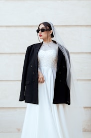 A person wearing a white wedding dress paired with a black jacket, sunglasses, and a veil, against a light-colored background. The dress is adorned with intricate patterns on the bodice.