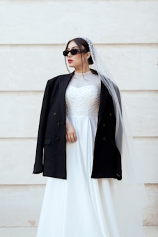 A person wearing a white wedding dress paired with a black jacket, sunglasses, and a veil, against a light-colored background. The dress is adorned with intricate patterns on the bodice.