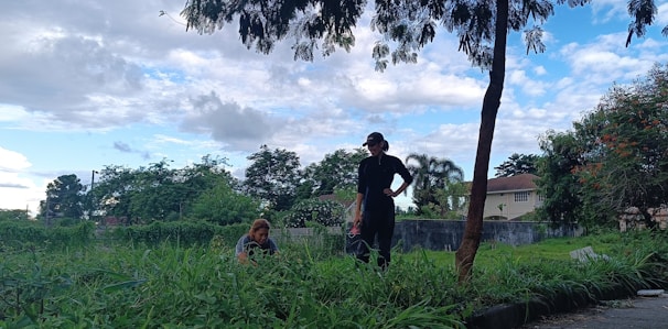 Two people are in a grassy garden area under a tree with dense foliage. One person is standing and seems to be overseeing or instructing, while the other is bent down, possibly working on the plants. The background includes green bushes, a few houses, and a partly cloudy sky.