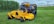 A yellow commercial lawn mower is parked on a well-maintained grassy field. In the background, there is a row of tall green corn plants and a metal guardrail separating the field from an industrial area. The sky is overcast with gray clouds.