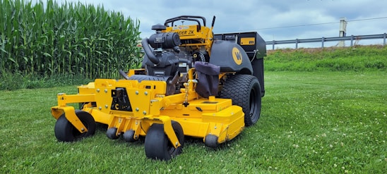 A yellow commercial lawn mower is parked on a well-maintained grassy field. In the background, there is a row of tall green corn plants and a metal guardrail separating the field from an industrial area. The sky is overcast with gray clouds.