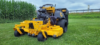 A yellow commercial lawn mower is parked on a well-maintained grassy field. In the background, there is a row of tall green corn plants and a metal guardrail separating the field from an industrial area. The sky is overcast with gray clouds.