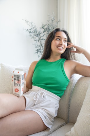 A woman sitting comfortably on a couch, wearing a green tank top and white shorts. She holds a carton labeled 'Boxed Water is Even Better' and looks to the side with a relaxed smile. The room has soft, natural lighting and a potted plant is visible in the background.