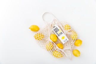 A white mesh bag holds several bright yellow lemons and a carton of boxed water. The tag on the carton states 'Boxed Water Is Better'. The objects are set against a clean, minimalistic background.
