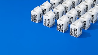 A neatly arranged group of cartons labeled 'Boxed Water' set against a vibrant blue background. The cartons are predominantly white with bold black text and positioned in orderly rows, creating a striking visual contrast.