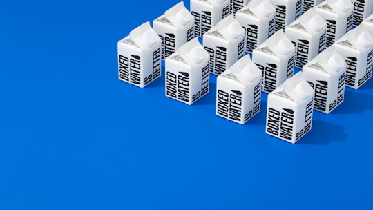 A neatly arranged group of cartons labeled 'Boxed Water' set against a vibrant blue background. The cartons are predominantly white with bold black text and positioned in orderly rows, creating a striking visual contrast.