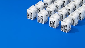 A neatly arranged group of cartons labeled 'Boxed Water' set against a vibrant blue background. The cartons are predominantly white with bold black text and positioned in orderly rows, creating a striking visual contrast.
