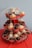 A tiered cupcake stand holds multiple cupcakes with both chocolate and vanilla frosting, decorated with colorful sprinkles. The cupcakes are arranged in red polka dot wrappers. A large number six candle and a 'Happy Birthday' decoration are prominently placed on the top tier. The stand is positioned on a red tablecloth against a light blue wall.