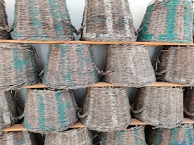 A collection of woven wicker baskets arranged on wooden shelves. The baskets have a natural color with a hint of green and beige patterns. They are stacked in rows, showcasing their tapered cylindrical shape.