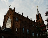 A large, ornate brick church with a Gothic architectural style. The building features tall spires, pointed arches, and large stained glass windows. A prominent cross sits atop one of the spires. The sky is partially cloudy, with some floral elements visible nearby.
