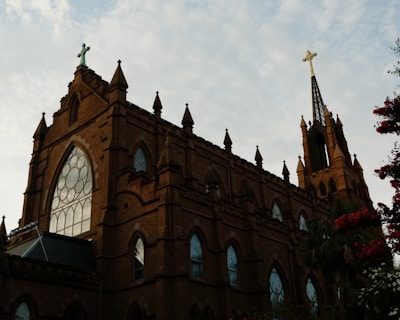 A large, ornate brick church with a Gothic architectural style. The building features tall spires, pointed arches, and large stained glass windows. A prominent cross sits atop one of the spires. The sky is partially cloudy, with some floral elements visible nearby.