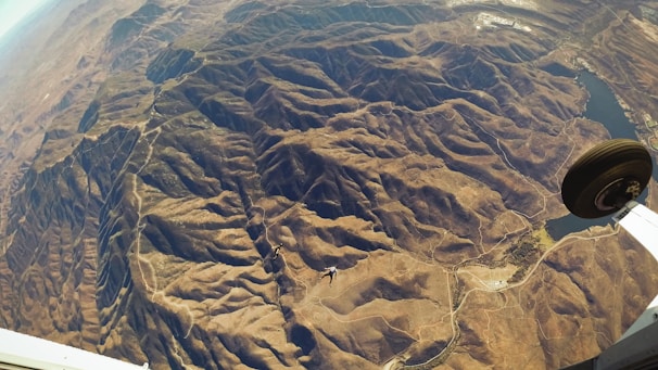 An aerial view of a mountainous landscape with rugged terrain, valleys, and scattered roads. A plane's landing gear can be seen on the right side of the image, and a small body of water is visible towards the top right. The overall landscape appears dry and arid.