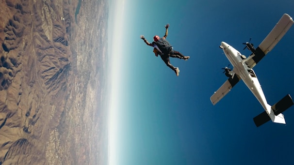 A skydiver wearing a red helmet is in freefall beside a small aircraft, high above a mountainous landscape. The sky is clear and blue, contrasting with the rugged terrain below.