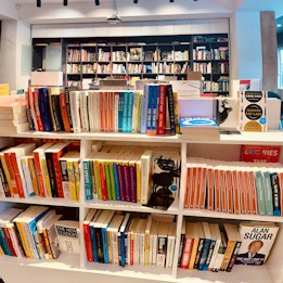 An indoor setting featuring a bookshelf filled with a variety of books. The shelves are neatly organized, displaying numerous book covers, some of which are bestsellers. The background includes additional bookshelves filled with more books, suggesting a library or bookstore environment.
