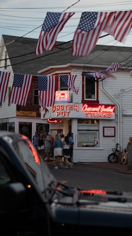 A street scene featuring a restaurant called Lobster Pot, which is characterized by neon signs. Several American flags hang above the street, and a group of people stand in front of the entrance. The scene includes a parked car in the foreground and a bicycle leaning against the building.