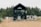 A modern tiny house with black paneling positioned in a natural setting surrounded by tall pine trees. A large glass door is the focal point, with two gray chairs on a wooden deck in front of it. To the right, a trailer or RV is visible, suggesting the location is possibly a camping or retreat area. The foreground features sandy soil with sparse tufts of grass, contributing to a rustic outdoor atmosphere.
