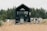 A modern tiny house with black paneling positioned in a natural setting surrounded by tall pine trees. A large glass door is the focal point, with two gray chairs on a wooden deck in front of it. To the right, a trailer or RV is visible, suggesting the location is possibly a camping or retreat area. The foreground features sandy soil with sparse tufts of grass, contributing to a rustic outdoor atmosphere.