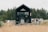 A modern tiny house with black paneling positioned in a natural setting surrounded by tall pine trees. A large glass door is the focal point, with two gray chairs on a wooden deck in front of it. To the right, a trailer or RV is visible, suggesting the location is possibly a camping or retreat area. The foreground features sandy soil with sparse tufts of grass, contributing to a rustic outdoor atmosphere.