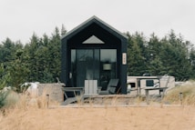 A modern tiny house with black paneling positioned in a natural setting surrounded by tall pine trees. A large glass door is the focal point, with two gray chairs on a wooden deck in front of it. To the right, a trailer or RV is visible, suggesting the location is possibly a camping or retreat area. The foreground features sandy soil with sparse tufts of grass, contributing to a rustic outdoor atmosphere.