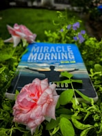 A book titled 'Miracle Morning' lies among lush green foliage and blooming flowers. The cover features a silhouette of a person with open arms against a calm sky. Dew-kissed pink roses and small purple flowers add to the serene setting.