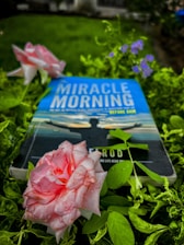 A book titled 'Miracle Morning' lies among lush green foliage and blooming flowers. The cover features a silhouette of a person with open arms against a calm sky. Dew-kissed pink roses and small purple flowers add to the serene setting.