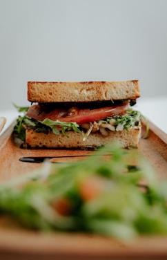A delicious sandwich layered with fresh tomatoes, leafy greens, and other fillings is positioned on a wooden serving tray. The foreground shows a blurred salad, creating a contrast between it and the focused sandwich.