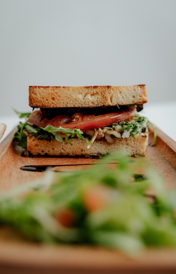 A delicious sandwich layered with fresh tomatoes, leafy greens, and other fillings is positioned on a wooden serving tray. The foreground shows a blurred salad, creating a contrast between it and the focused sandwich.