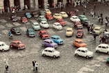 A collection of vintage cars in a variety of colors is parked on a cobblestone square, surrounded by a small crowd of people. The scene appears to be a gathering or exhibition, with onlookers taking photos and observing the vehicles.