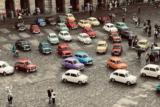 A collection of vintage cars in a variety of colors is parked on a cobblestone square, surrounded by a small crowd of people. The scene appears to be a gathering or exhibition, with onlookers taking photos and observing the vehicles.