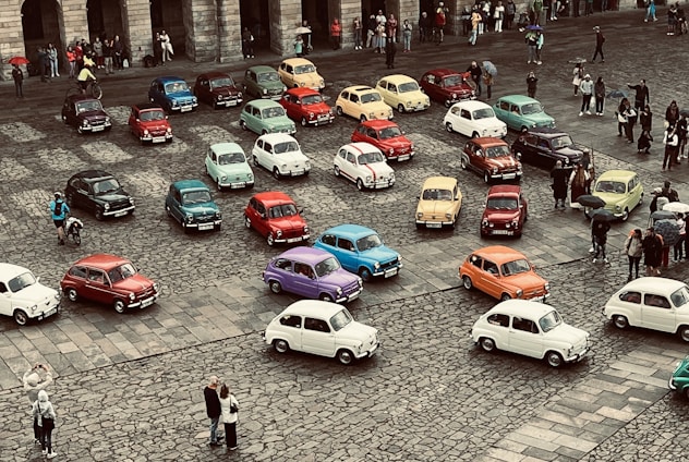 A collection of vintage cars in a variety of colors is parked on a cobblestone square, surrounded by a small crowd of people. The scene appears to be a gathering or exhibition, with onlookers taking photos and observing the vehicles.