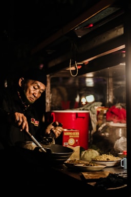 A man is cooking in a dimly lit street food stall. He is focused on stirring food in a pan with a ladle. In the background, items such as a red container, a box labeled 'Khong Guan Biscuits,' some plates, and vegetables like cabbage and beans can be seen.