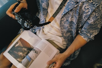 A person is sitting on a couch wearing a blue and white patterned shirt, holding a black camera with both hands. An open magazine rests on their lap, displaying photographs and text.