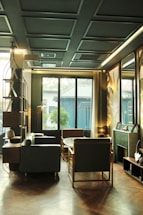 A cozy, well-decorated room featuring modern furniture, including a sofa, chairs, and a round table. The room has a patterned ceiling and large windows allowing natural light. The atmosphere is elegant with decorative items on shelves and a soft color palette.