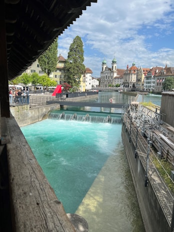 A scenic view of a river with bright turquoise water cascading over a small dam. Architectural buildings with red and green rooftops are visible in the background, along with a church featuring distinctive green domes. Lush green trees and a clear blue sky complete the picturesque urban landscape.