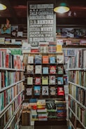A well-organized bookstore shelf filled with a variety of books. The central section has a display with books displayed with covers facing forward, showcasing titles from different genres and authors. Above the books, there is a large sign featuring the names of famous authors in bold, such as William Shakespeare and Jane Austen. The surrounding shelves are filled with more books arranged side-by-side.