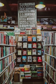 A well-organized bookstore shelf filled with a variety of books. The central section has a display with books displayed with covers facing forward, showcasing titles from different genres and authors. Above the books, there is a large sign featuring the names of famous authors in bold, such as William Shakespeare and Jane Austen. The surrounding shelves are filled with more books arranged side-by-side.