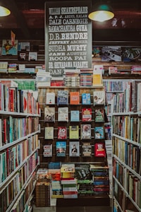 A well-organized bookstore shelf filled with a variety of books. The central section has a display with books displayed with covers facing forward, showcasing titles from different genres and authors. Above the books, there is a large sign featuring the names of famous authors in bold, such as William Shakespeare and Jane Austen. The surrounding shelves are filled with more books arranged side-by-side.