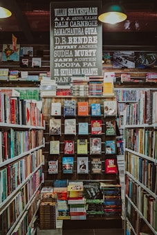 A well-organized bookstore shelf filled with a variety of books. The central section has a display with books displayed with covers facing forward, showcasing titles from different genres and authors. Above the books, there is a large sign featuring the names of famous authors in bold, such as William Shakespeare and Jane Austen. The surrounding shelves are filled with more books arranged side-by-side.