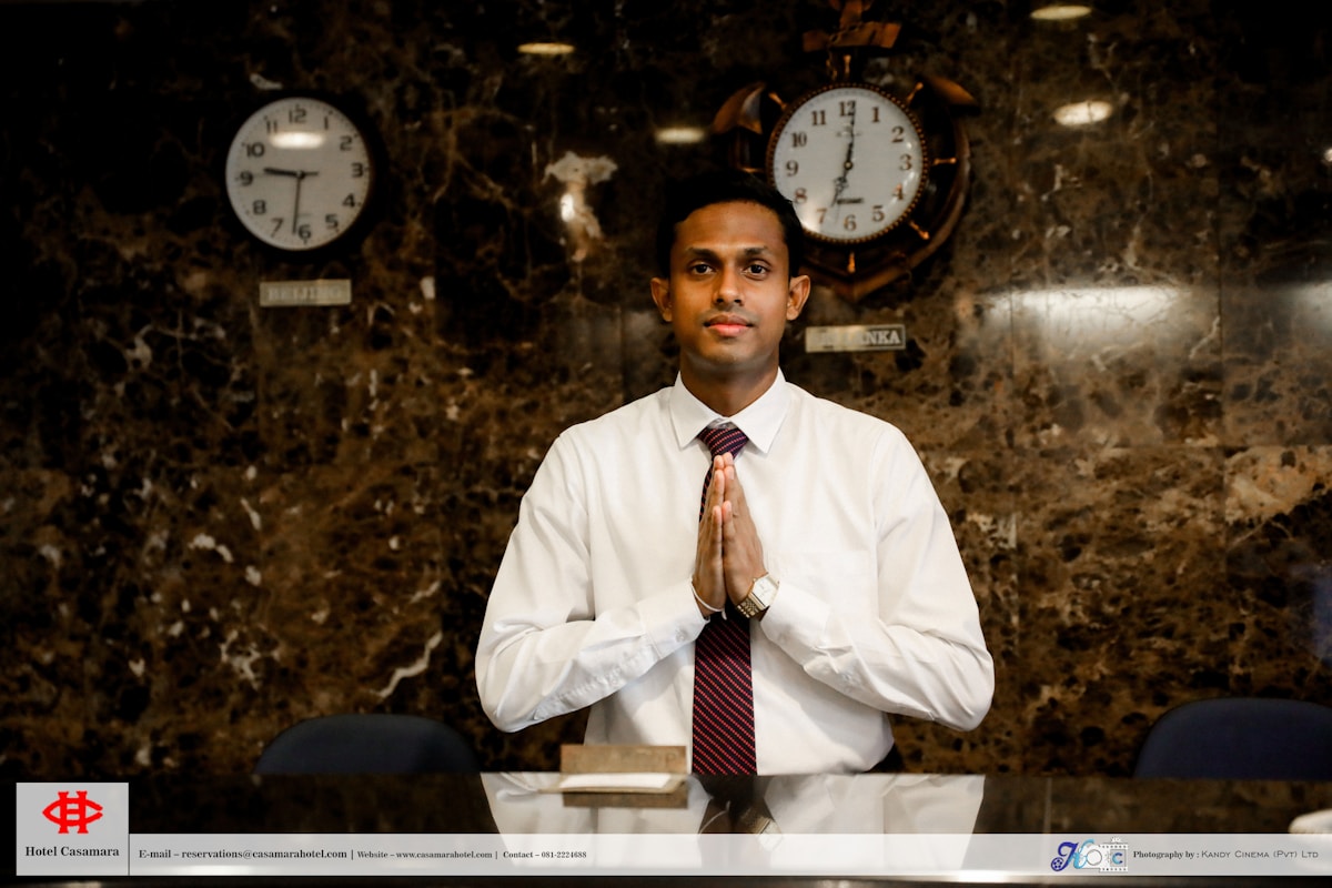 Hotel receptionist in a white shirt and red tie, hands clasped, at a marble desk with clocks behind.